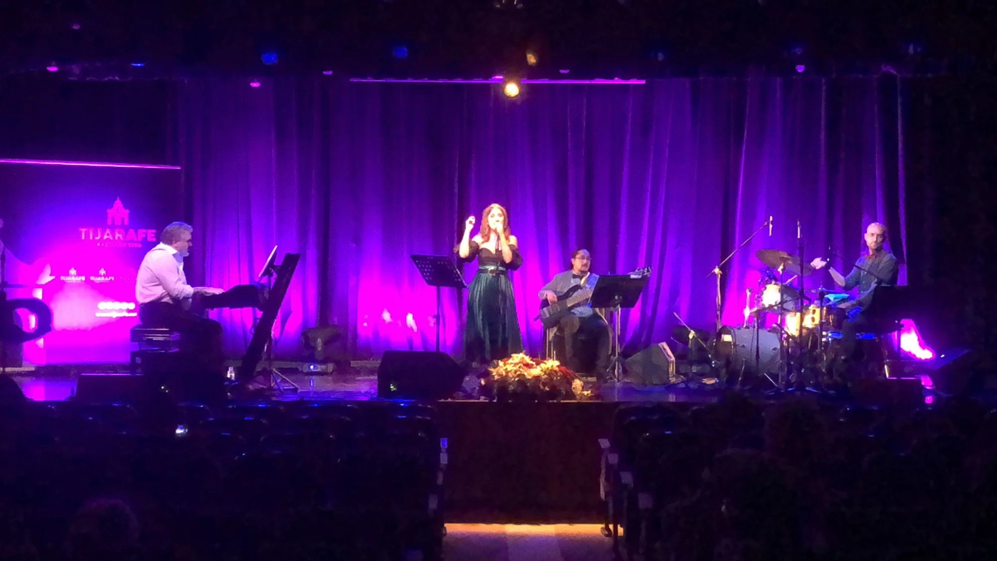 Concierto en sala con iluminación morada: piano, voz, bajo y batería en el escenario